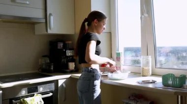 Güneşli bir mutfakta, genç bir kadın cam kenarında dururken malzemeleri bir kaseye karıştırıyor. Sıcak ışık, yemeklerine odaklanırken rahat bir atmosfer yaratıyor..