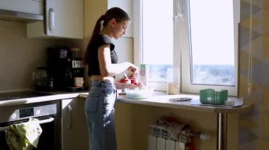 Parlak bir mutfakta, güneş ışığı pencereden içeri girerken, genç bir kadın kahvaltı için malzemeleri karıştırıyor. Yemek yaparken aşçılık yeteneklerini sergiliyor..