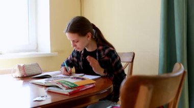Genç bir kız, kitapları ve kırtasiyelerle çevrili ahşap bir masada derslerine konsantre oluyor. Güneş ışığı pencereden içeri akar ve sıcak bir atmosfer yaratır..