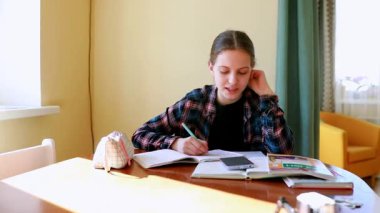 Kendini işine adamış bir öğrenci ahşap bir masada derslerine odaklanır. Ders kitapları ve hesap makinesiyle çevriliyken bir deftere not alıyor, hepsi de doğal ışıkla aydınlatılıyor..