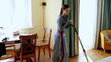 Bir insan evini temizlemekle meşgul, iyi aydınlatılmış bir odada vakum kullanıyor. Mekan rahat, basit mobilyalar ve parlak renkler sergileniyor, temiz bir çevreyi yansıtıyor..
