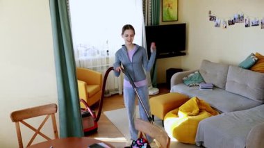 Genç bir insan, yumuşak mobilya ve dekorla dolu rahat bir oturma odasını süpürüyor. Güneş pencereden parlıyor, davetkar bir atmosfer yaratıyor..