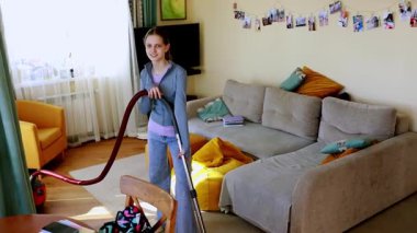 Bir kız zevkle oturma odasını elektrikli süpürgeyle temizler. Odada rahat bir kanepe, renkli yastıklar ve pencereden süzülen güneş ışığı var..