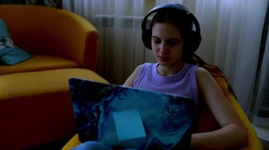 Genç bir kadın parlak bir kanepede rahatça oturuyor, kulaklık takıyor ve dikkatle dizüstü bilgisayarına odaklanıyor. Konforlu ortam yumuşak mobilyalar ve sıcak ışığa sahiptir..