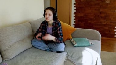 Genç bir kadın sıcak bir oturma odasındaki kanepede bağdaş kurup kulaklık takmış oyun oynuyor. Güneş ışığı pencereden akar ve rahatlatıcı bir atmosfer yaratır..