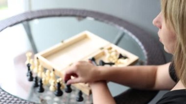 Bir kadın terasta oturur ve tahta bir tahtanın üzerinde satranç oynar. Oyuna odaklanmış ve stratejik hamleler yapıyor..