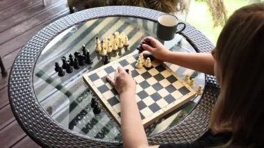 Teras masasında cam kapaklı satranç oynayan bir kadın. Dışarıda güneşin altında oturuyor ve oyununa konsantre oluyor..