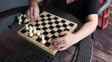 Terasta oturan bir kadın satranç oynuyor. Satranç tahtası masanın ortasında, ve kadın da siyah bir gömlek giyiyor. Parlak, güneşli bir gün..