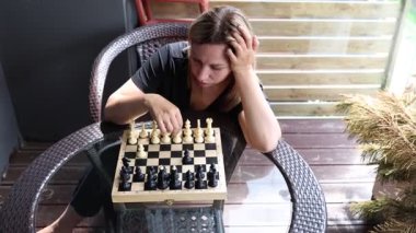 Bir kadın güneşli terasta satranç oynuyor. Hasır bir sandalyede oturuyor, bir sonraki hamlesini planlarken konsantrasyonunun derinliklerinde. Satranç tahtası cam bir masaya kurulmuş ve güneş...