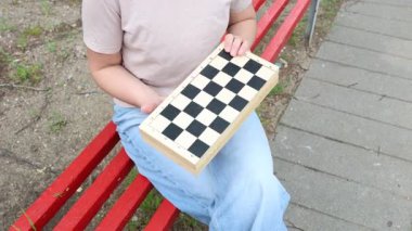 Kırmızı parktaki bankta oturan ve dama oynayan bir kadın. Tahtada bir parçayı hareket ettiriyor, oyuna konsantre oluyor..