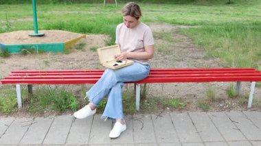 Parkta kırmızı bir bankta oturan bir kadın. Kamera dışında biriyle dama oynuyordur. Bankta ağaçların gölgesi var ve kadın günlük giysiler giyiyor..
