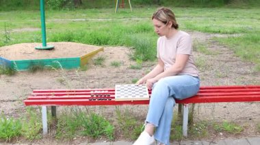 Bu video parkta kırmızı bir bankta dama oynayan bir kadını gösteriyor. O bankta oturuyor ve elinde bir dama tahtası tutuyor. Video muhtemelen gündüz çekilmiş..