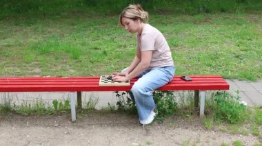 Parkta kırmızı bir bankta oturan ve dama oynayan bir kadın. Oyuna odaklanmış ve eğleniyor gibi görünüyor. Güneş parlıyor ve gün aydınlık.