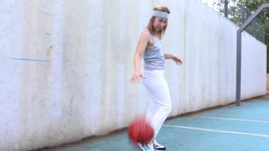 Bu video, genç beyaz bir kadının bir spor sahasında basketbol antrenmanı yaptığını gösteriyor. Beyaz pantolon ve beyaz bir atlet giyiyor. Topu sürüyor ve sahanın etrafında dönüyor..