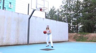 Saç bandı takan genç bir kadın spor sahasında basketbol oynuyor. Top sürüyor ve yeteneklerini geliştiriyor. Video, güneşli bir günde gündüz çekiliyor..