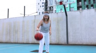 Bu video açık hava sahasında basketbol oynayan beyaz bir kadını gösteriyor. O, atletik giysiler giyiyor ve topu sürüyor. Saray yüksek bir bina ve beton bir duvarla çevrili.