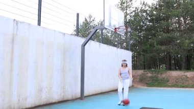Bu video, gün içinde spor sahasında basketbol oynayan beyaz bir kadını gösteriyor. Beyaz elbiseler giyiyor ve potaya doğru basket oynarken görülüyor. Video bir filmde çekildi.