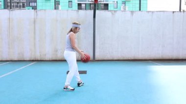 Bu video beyaz eşofmanlı ve gri kolsuz tişörtlü genç bir kadını mavi kortta basketbol antrenmanı yaparken gösteriyor. Ayak hakimiyeti ve top tutuş becerileri üzerinde çalışıyor..