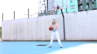 Bu video, spor sahasında basketbol oynayan beyaz elbiseli genç bir kadını gösteriyor. Güneş ışıl ışıl parlıyor ve o yeteneklerini geliştirirken eğleniyor..