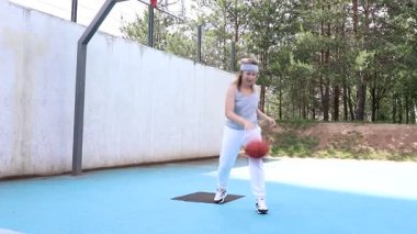 Beyaz pantolonlu ve gri atletli genç bir kadın güneşli bir günde basketbol antrenmanı yapıyor. Açık havada basketbol sahası kullanıyor, arka planda bir halka var. Top sürüyor ve antrenman yapıyor.