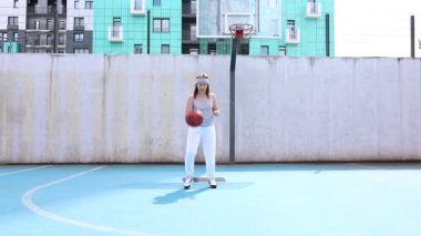 Beyaz pantolonlu ve gri tişörtlü genç bir kadın basketbol yeteneklerini modern bir apartmanın önünde açık havada çalışıyor. Basketbol topunu önünde tutuyor ve odaklanmış görünüyor..