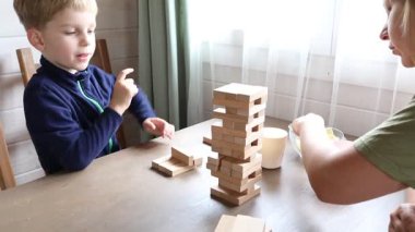 Bir anne ve oğlu mutfak masasında Jenga oynuyorlar. Genç çocuk dikkatlice bir blok çıkarırken oyuna odaklanmış durumda. Annesi onun karşısında oturur ve dikkatle izlerdi..