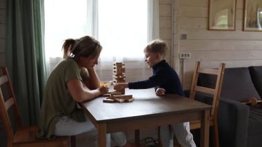 Bir anne ve oğlu mutfak masasında klasik Jenga oyunu oynuyorlar. Çocuk bir sandalyede oturuyor ve bir bloğa uzanıyor, anne onun yanında bir sandalyede oturuyor, dikkatle izliyor.