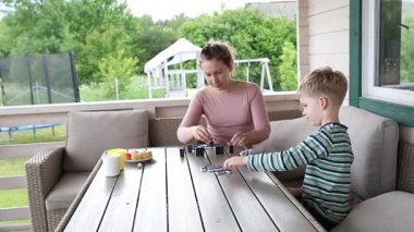 Bir anne ve iki küçük oğlu güneşli bir yaz gününde domino oynuyorlar. Tahta bir masa ve hasır sandalyelerle terasta oturuyorlar. Aile rahatlamış ve eğleniyor gibi görünüyor.
