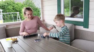 Bir anne ve oğlu bir yaz günü tahta terasta domino oynuyorlar. Çocuk hasır bir sandalyede oturuyor ve anne bir masada oturuyor. İkisi de gülümsüyor ve harika vakit geçiriyorlar.