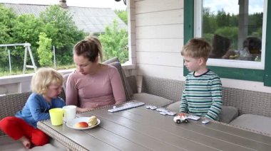Bir anne ve iki küçük oğlu terasta güneşli bir öğleden sonra birlikte domino oynuyorlar. Çocuklar ahşap bir masada otururken, anne karşılarında oturuyor, onların oynamasını izliyor..