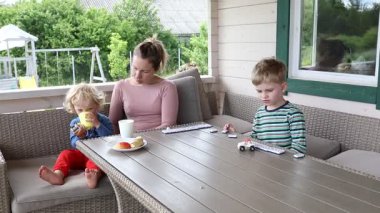 Bir anne ve iki küçük oğlu teraslarında rahatlatıcı bir öğleden sonranın tadını çıkarıyorlar. Küçük oğlu sarı bardaktan içerken onlar domino oynuyorlar. Birlikte çok eğleniyor gibi görünüyorlar..