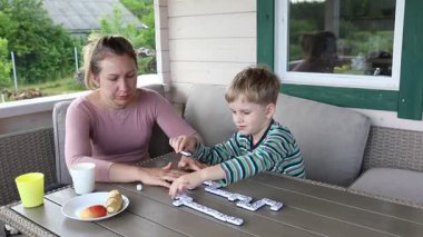 Bir anne ve iki küçük oğlu güneşli bir terasta domino oynuyorlar. Çocuklar anneleriyle bir masada oturuyorlar ve oynarken hepsi gülümsüyor ve gülüyorlar..