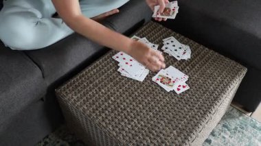 Açık mavi gömlekli bir kadın koltukta otururken hasır bir koltukta kağıt oynuyor. Kartlar kanepeye rastgele dizilmiş gibi görünüyor..