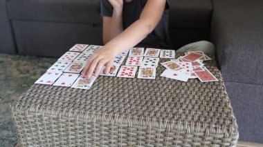 Bu video, oturma odasındaki hasır masada kağıt oynayan bir kadını gösteriyor. Kadın gri bir gömlek giyiyor..