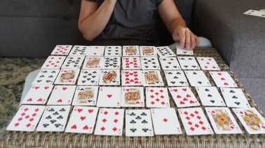 Oturma odasındaki masada kağıt oynayan bir kadın. Masa hasır bir masa örtüsüyle kaplı ve arka planda gri bir kanepe var. Kağıtları belli bir düzende hazırlıyor gibi görünüyor ama ne tür bir oyun oynadığı belli değil.