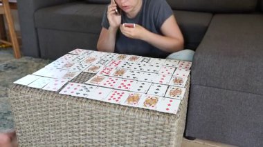 Bir kadın oturma odasındaki bir kanepede otururken hasır bir masanın üzerine iskambil kağıdı seriyor. Kartları sırayla koyuyor ve bir kart oyunu oynuyor gibi görünüyor..
