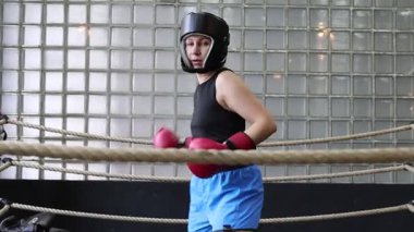Bu video spor salonunda boks antrenmanı yapan bir kadını gösteriyor. Siyah bir atlet, mavi şort, boks eldiveni ve koruyucu bir başlık giyiyor. Boks ringinde yumruk pratiği yapıyor..