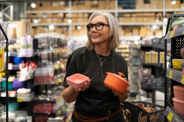 Olgun bir kadın, özenle seçtiği yemek takımları, dekoratif eşyalar ve bitkilerle organize edilmiş bir ev eşyaları süpermarketine göz atar ve bağımsız yaşam tarzını sergiler..