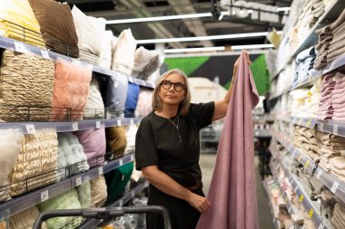 Modern bir perakende mağazasında renkli kumaşlara ve tekstil ürünlerine bakan şık bir olgun kadın. Kişisel zevklerini yansıtarak seçeneklerini dikkatlice değerlendiriyor..