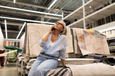 Bir kadın, ev eşyaları bölümünde kumaş ve dekoratif eşyaların arasında otururken düşünceli görünür. Bir telefon görüşmesi yapıyor, alışveriş tercihlerini düşünüyor..