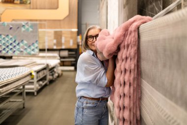 Bir kadın hipermarkette tekstil ürünlerine göz atar, çeşitli dekor ürünleriyle neşe içinde etkileşim kurar. Onun ifadesi alışverişin heyecanını ve ev eşyalarını keşfetmeyi yansıtıyor..