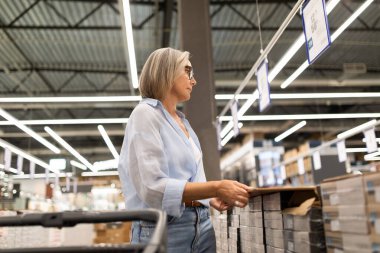 Canlı bir hipermarkette, bir kadın ürünleri çeşitli bölümlerde incelikle inceler, alışveriş deneyimindeki malzemelerle etkileşime girerken gerçek duygular gösterir..