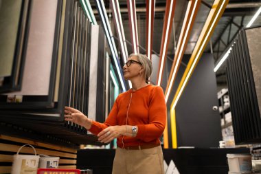 Orta yaşlı bir kadın, mutfak dekorasyonu ve bahçe malzemeleri gibi farklı bölümlerdeki ürünleri inceleyerek bir ev geliştirme mağazasına göz atıyor. Alışveriş yaparken vitrinlerle etkileşime giriyor..