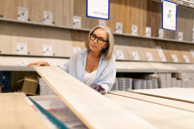 Orta yaşlı bir kadın, bir ev geliştirme mağazasının çeşitli bölümlerini karıştırıyor, ürünleri inceliyor ve el arabasına eşyalar yerleştiriyor. Atmosfer günlük alışveriş ve tüketici alışkanlıklarını yansıtıyor.