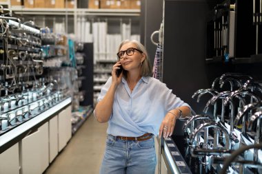 Bir kadın perakende mağazasında ev eşyaları karıştırıyor, telefonuyla konuşuyor, modern mutfak aletlerini dikkatlice seçerken, hakiki duygularını sergiliyor ve ürünlerle etkileşimde bulunuyor..