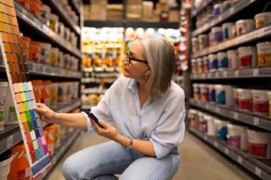 Orta yaşlı bir kadın kalabalık bir inşaat hipermarketinde çok çeşitli boya örnekleri keşfediyor, evi için aktif olarak renk seçiyor. Yakınlarda bir alışveriş arabası..