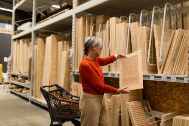 Ev geliştirme mağazasında, orta yaşlı bir kadın vitrinlerle etkileşim halinde olan ve alışveriş arabasını ev ve bahçe projeleri için seçenekler ile dolduran dekor bölümündeki eşyaları inceliyor..