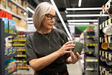 Bir kadın hipermarkette yeşil bir kahve fincanını inceler, çeşitli eşyalarla çevrilidir. Mutfak gereçleri bölümündeki ürünlerle etkileşime geçiyor ve alışveriş deneyimini gösteriyor..