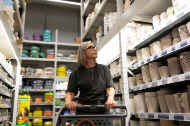 Canlı bir hipermarkette, bir kadın düşünceli bir şekilde ev eşyaları, dekorlar ve bitkilerle dolu koridorlarda dolaşır, gerçek duygular gösterir ve ürünlerle etkileşim kurar..