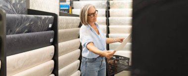 Orta yaşlı bir kadın, alışveriş deneyiminin bir parçası olarak mağaza bölümlerini karıştırıyor, eşyaları aktif olarak kontrol ediyor ve alışveriş arabasında seçimler yapıyor..